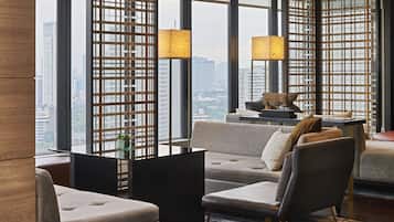 A modern hotel room with a city view, a sofa, chairs, a small table with a lamp, and decorative items.