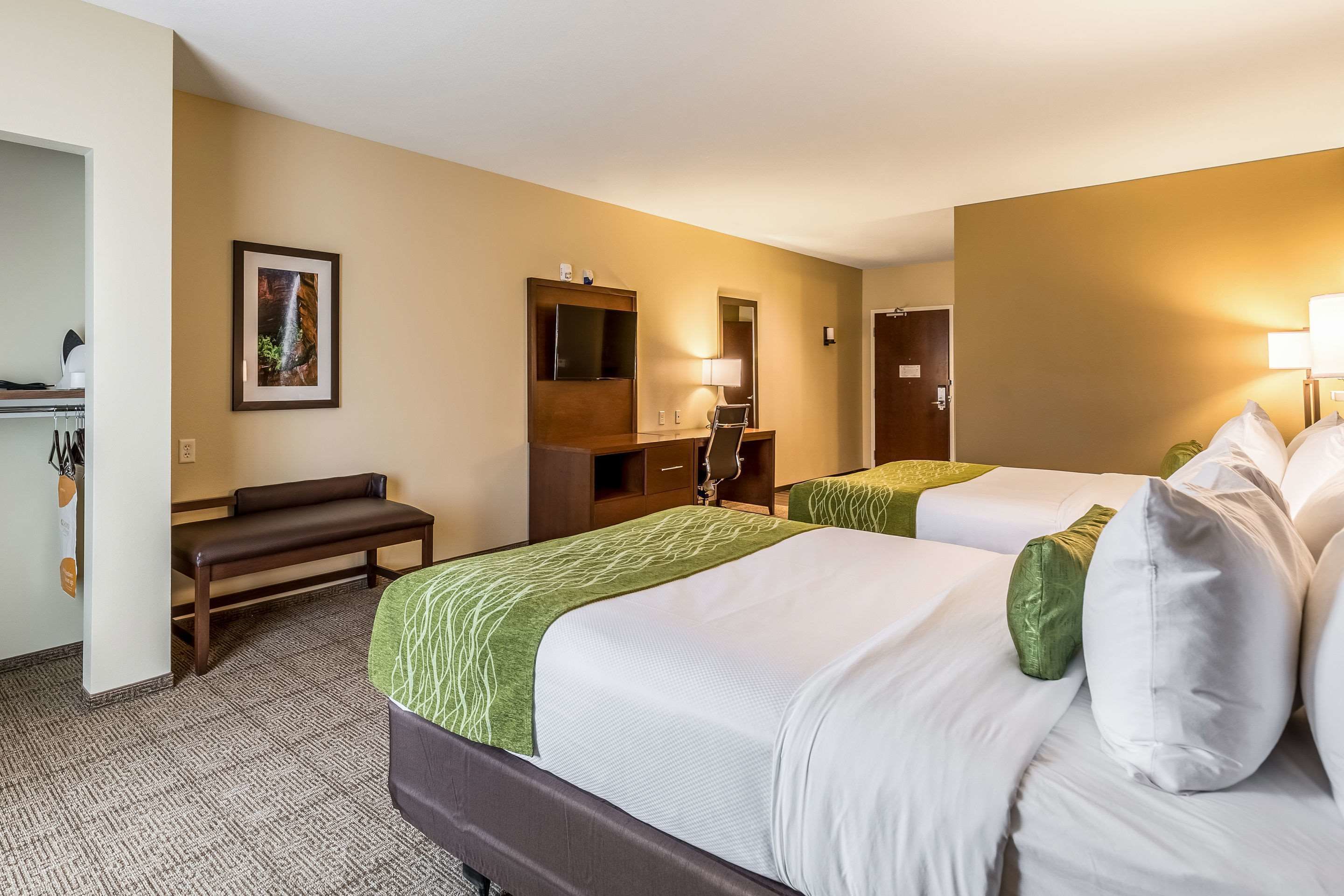 A hotel room with two beds, a bench, a desk with a lamp, a mirror, and a framed picture on the wall.