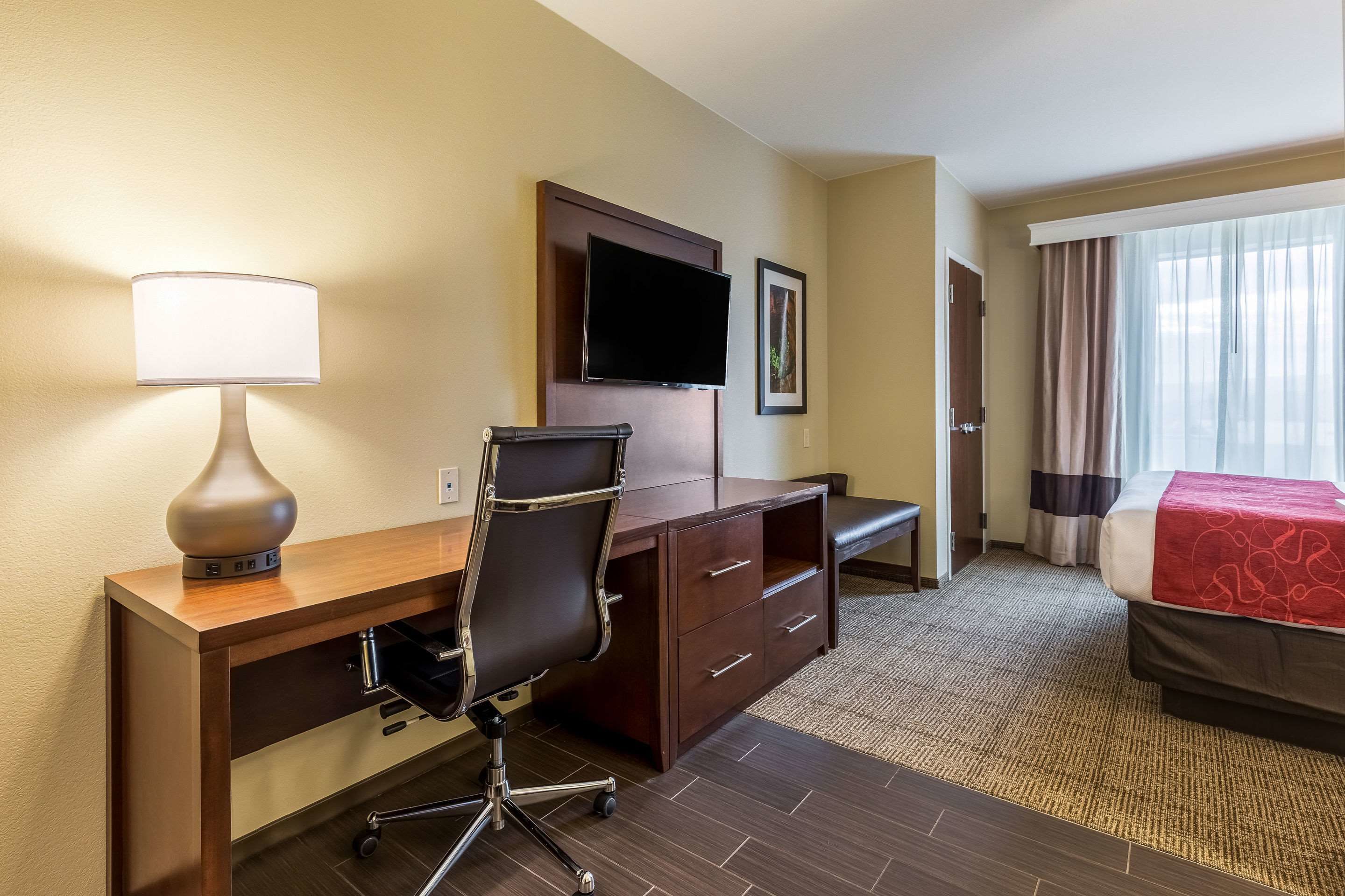 Een hotelkamer met een bureau, stoel, televisie, bed en groot raam.
