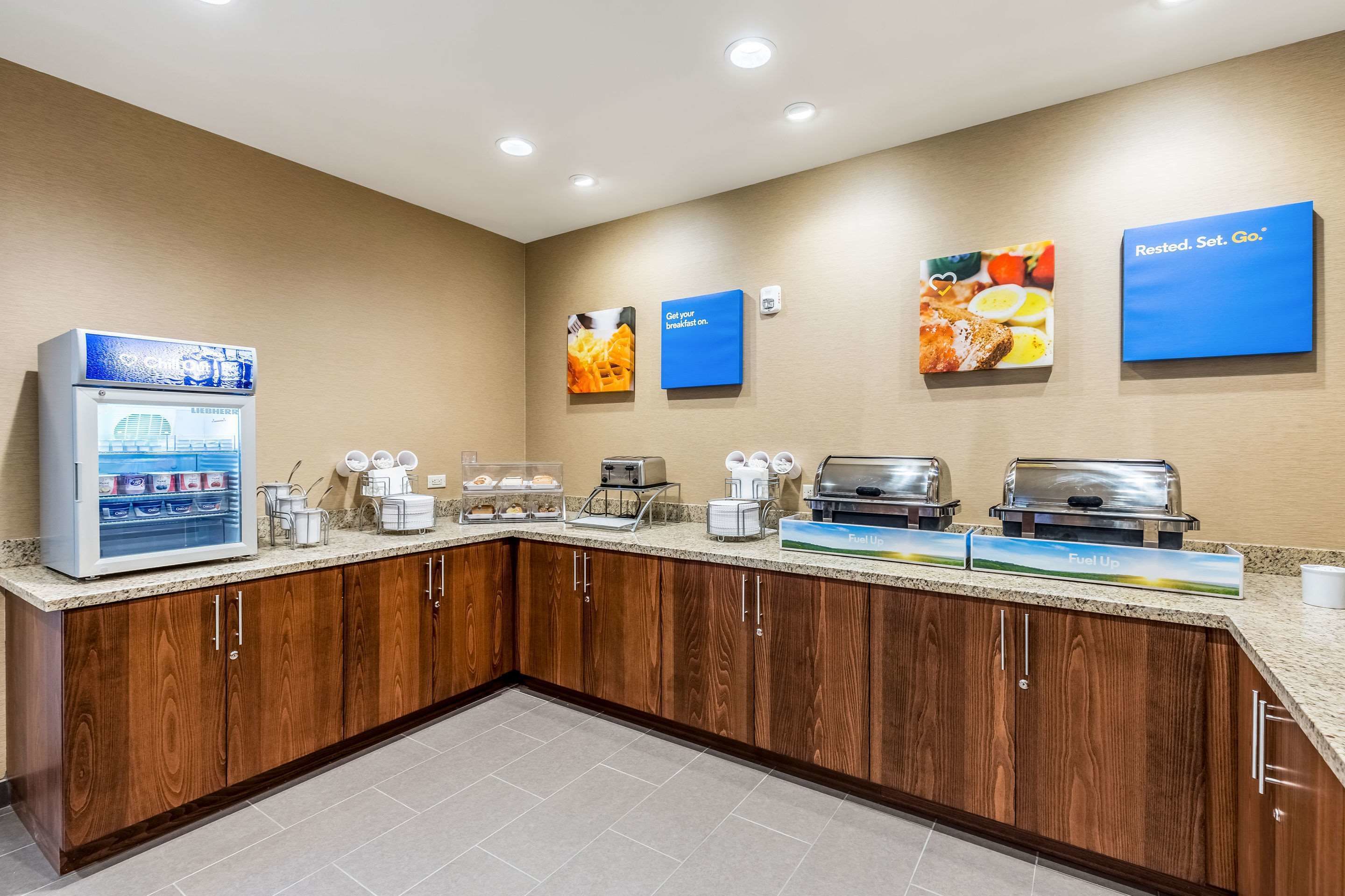 A hotel breakfast area with a self-service station, coffee makers, and a refrigerator.
