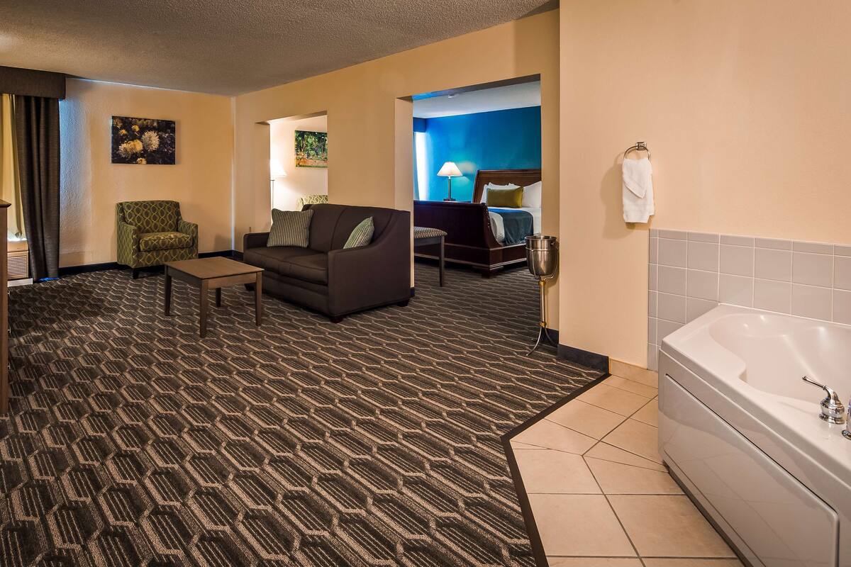 A hotel room with a patterned carpet, a sofa, a small table, a bed with a blue headboard, a bathroom with a bathtub, and a television.