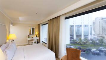 A hotel room with a bed, a desk, a chair, and a window with a view of a cityscape and parked cars.