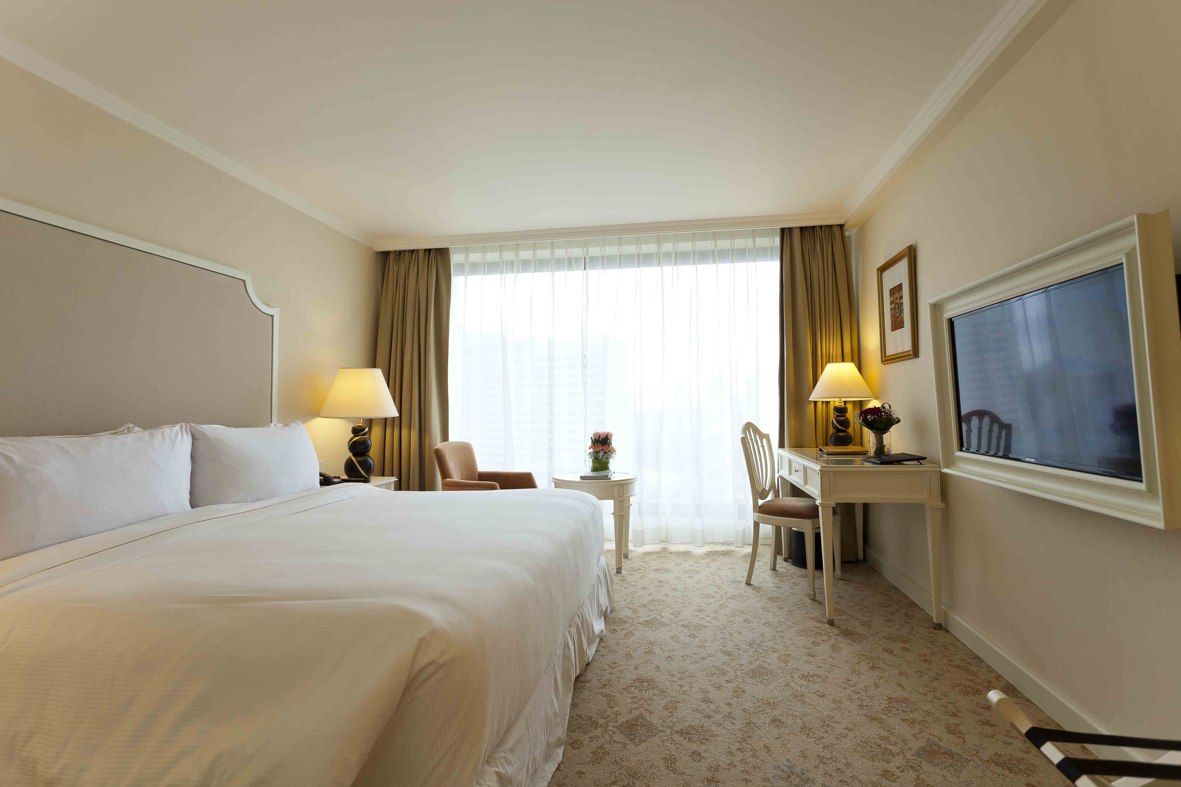 A hotel room with a large bed, a desk with a chair, a small table with a lamp, and a large window with curtains.