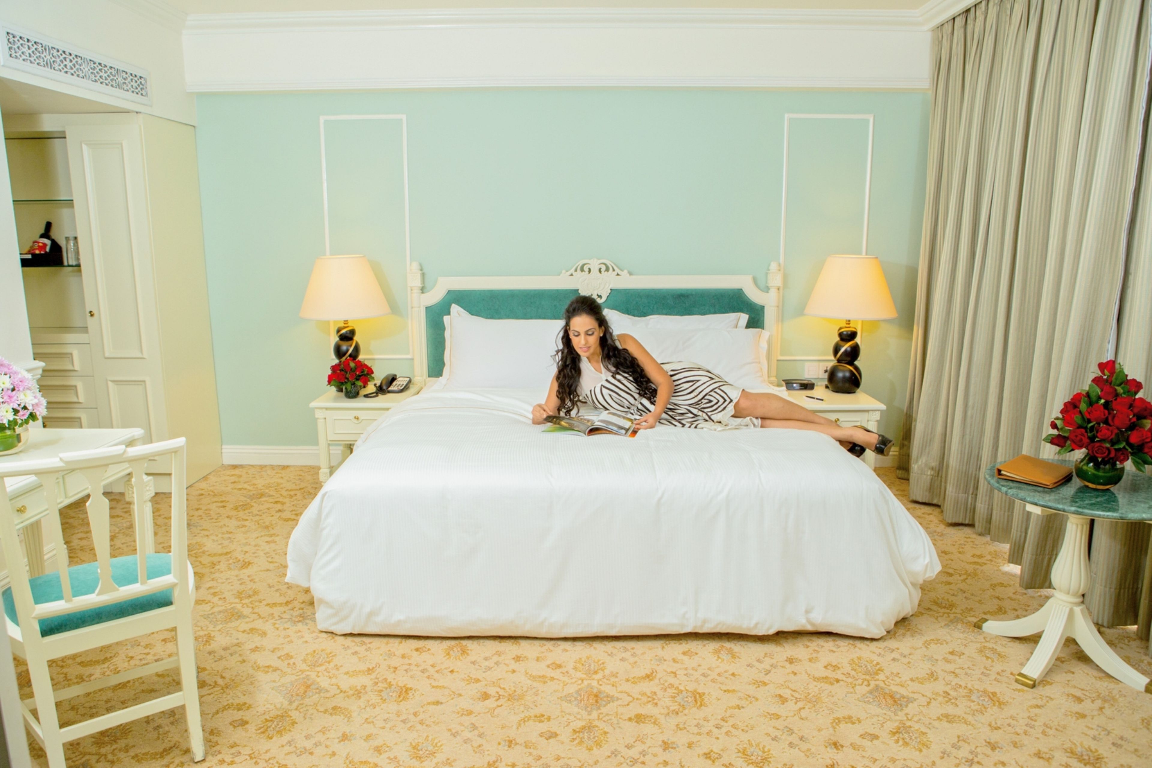 Habitación de hotel con cama, dos mesitas de noche con lámparas, una silla, una mesita con un jarrón de flores y una puerta que da a un armario.