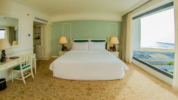 A hotel room with a large bed, two bedside tables with lamps, a desk with a chair, and a view of the beach.