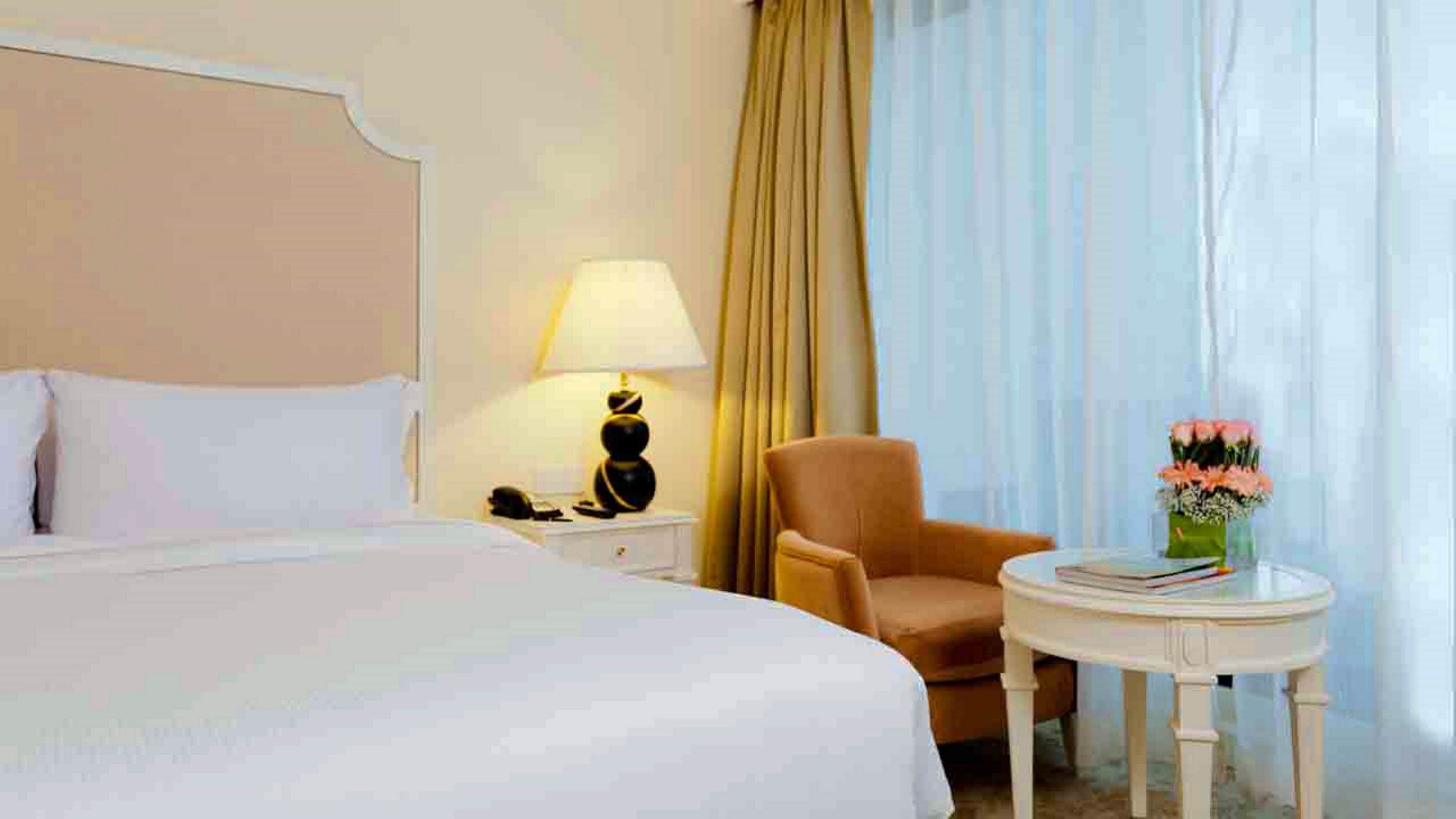 A hotel room with a large bed, a chair, a nightstand with a lamp, and a small table with a vase of flowers.