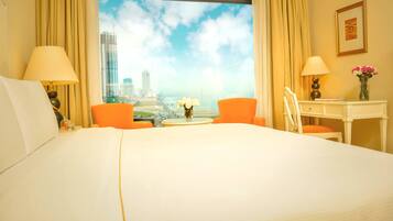 Ein Hotelzimmer mit einem großen Bett, zwei Sesseln, einem Schreibtisch und Blick auf eine Stadtlandschaft durch ein Fenster.