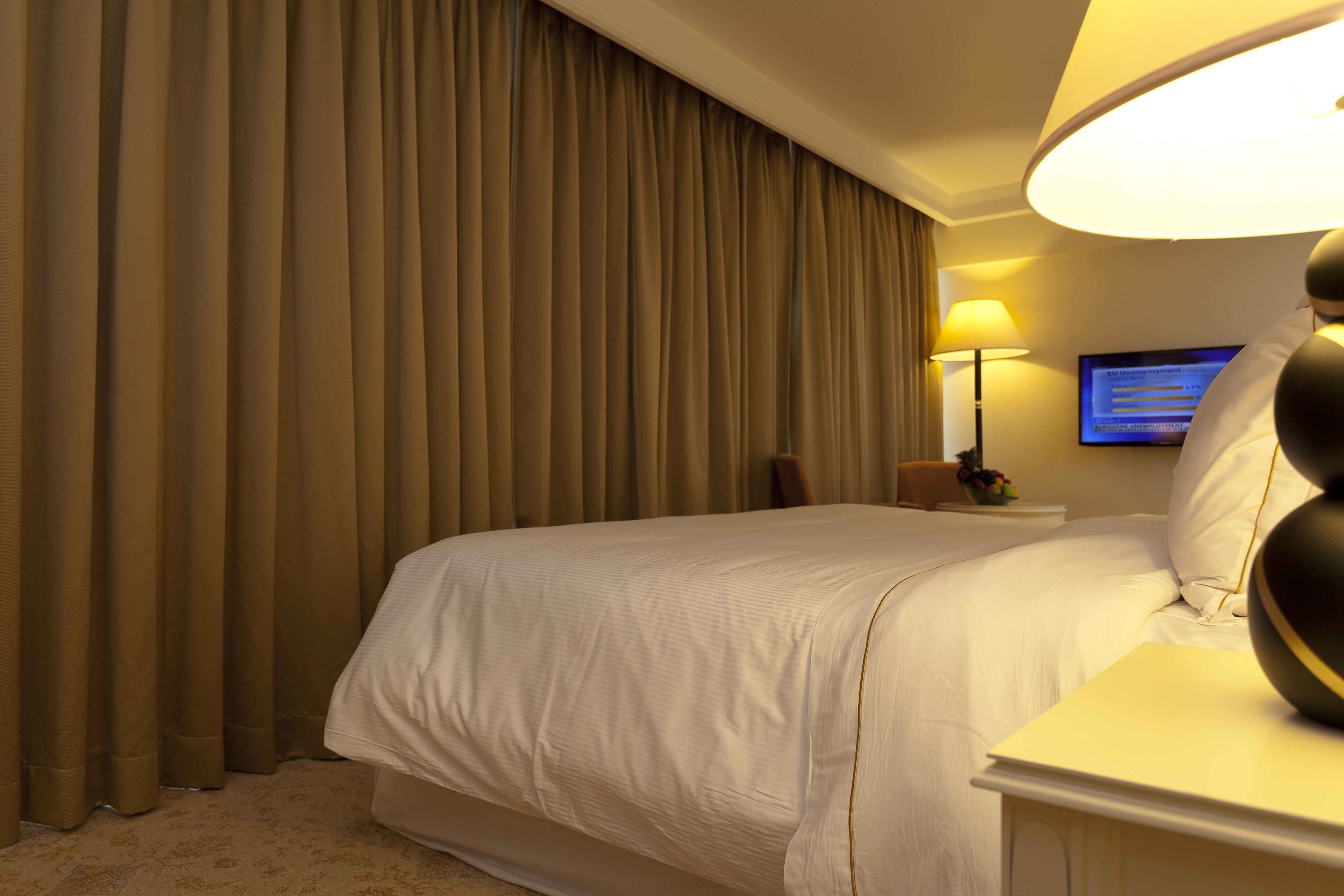 Una habitación de hotel con una cama grande, una mesita de noche con lámpara, un televisor en la pared y una ventana con cortinas.