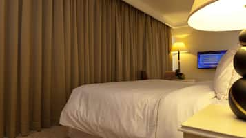 Una habitación de hotel con una cama grande, una mesita de noche con lámpara, un televisor en la pared y una ventana con cortinas.