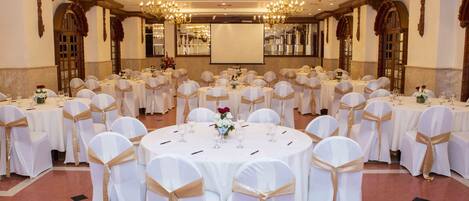 Un salón de banquetes con mesas redondas, manteles blancos y fajas doradas, equipado con candelabros y una pantalla grande.
