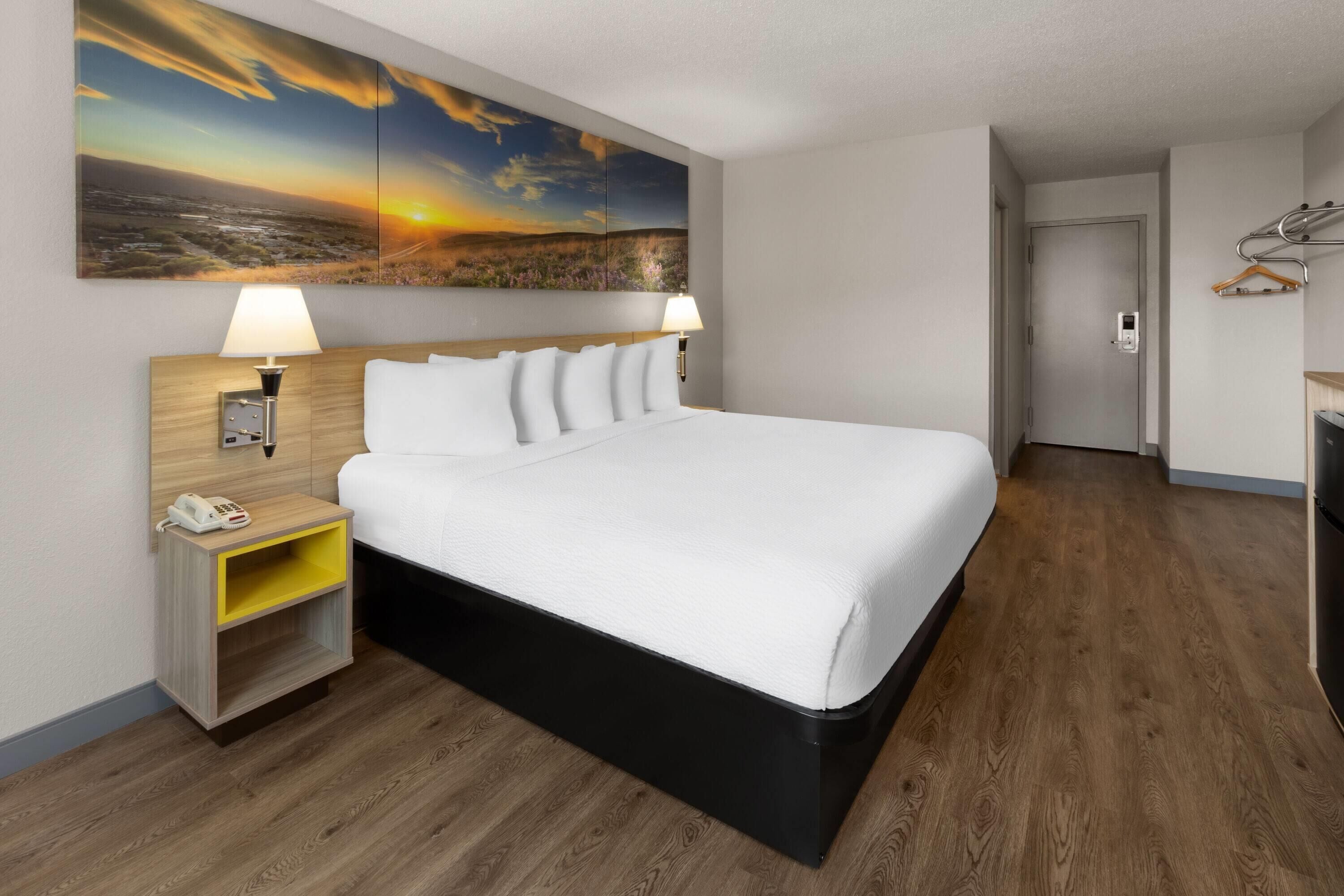 Ein Hotelzimmer mit einem großen Bett, zwei Nachttischlampen, einem Nachttisch mit Telefon, einem Holzboden und einem Sonnenuntergangsbild an der Wand.
