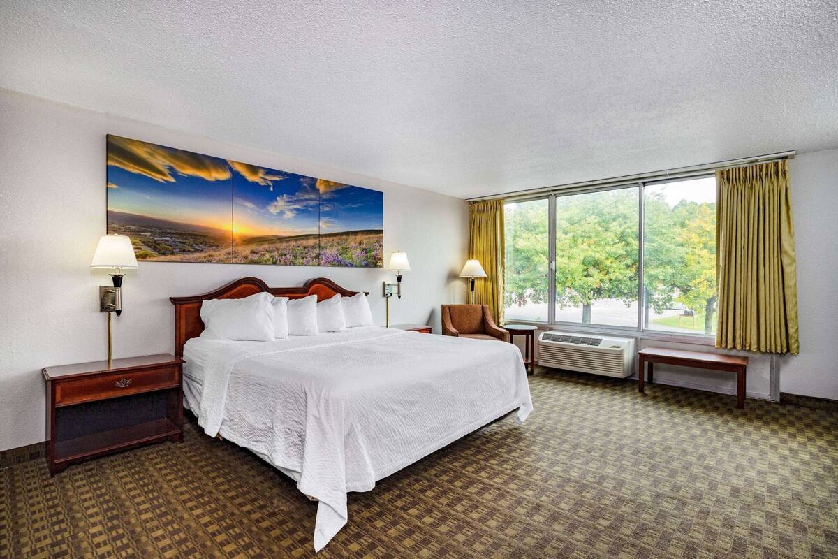 Ein Hotelzimmer mit einem großen Bett, zwei Nachttischen mit Lampen, einem Sessel, einem Fenster mit Vorhängen und einem Bild an der Wand.
