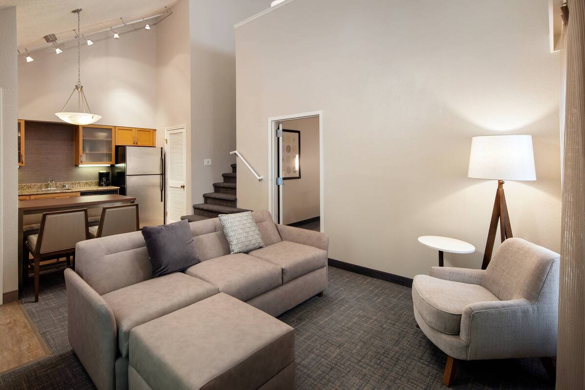 A modern living room with a sectional sofa, a small round table, a lamp, and a kitchen area in the background.