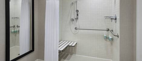 A walk-in shower with a white tiled floor, a showerhead, a handheld shower, a grab bar, and a bench.