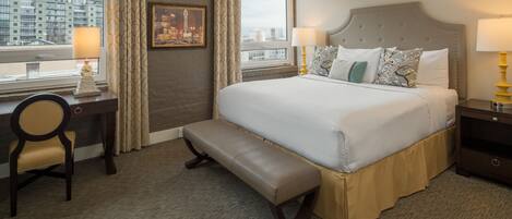 A hotel room with a large bed, a bench, a desk, and a view of the city.