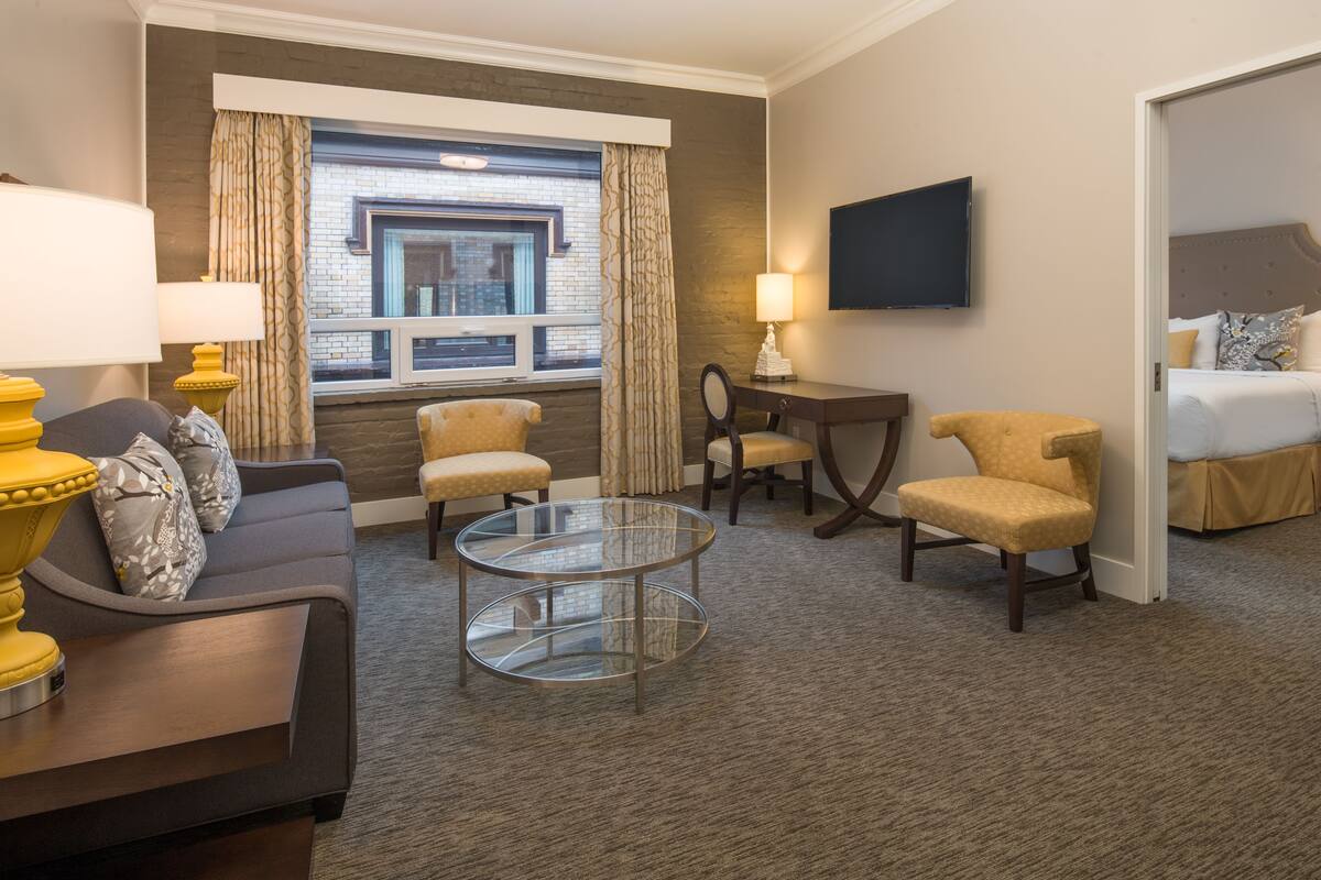 A hotel room with a sofa, armchairs, a glass coffee table, a desk, and a TV.