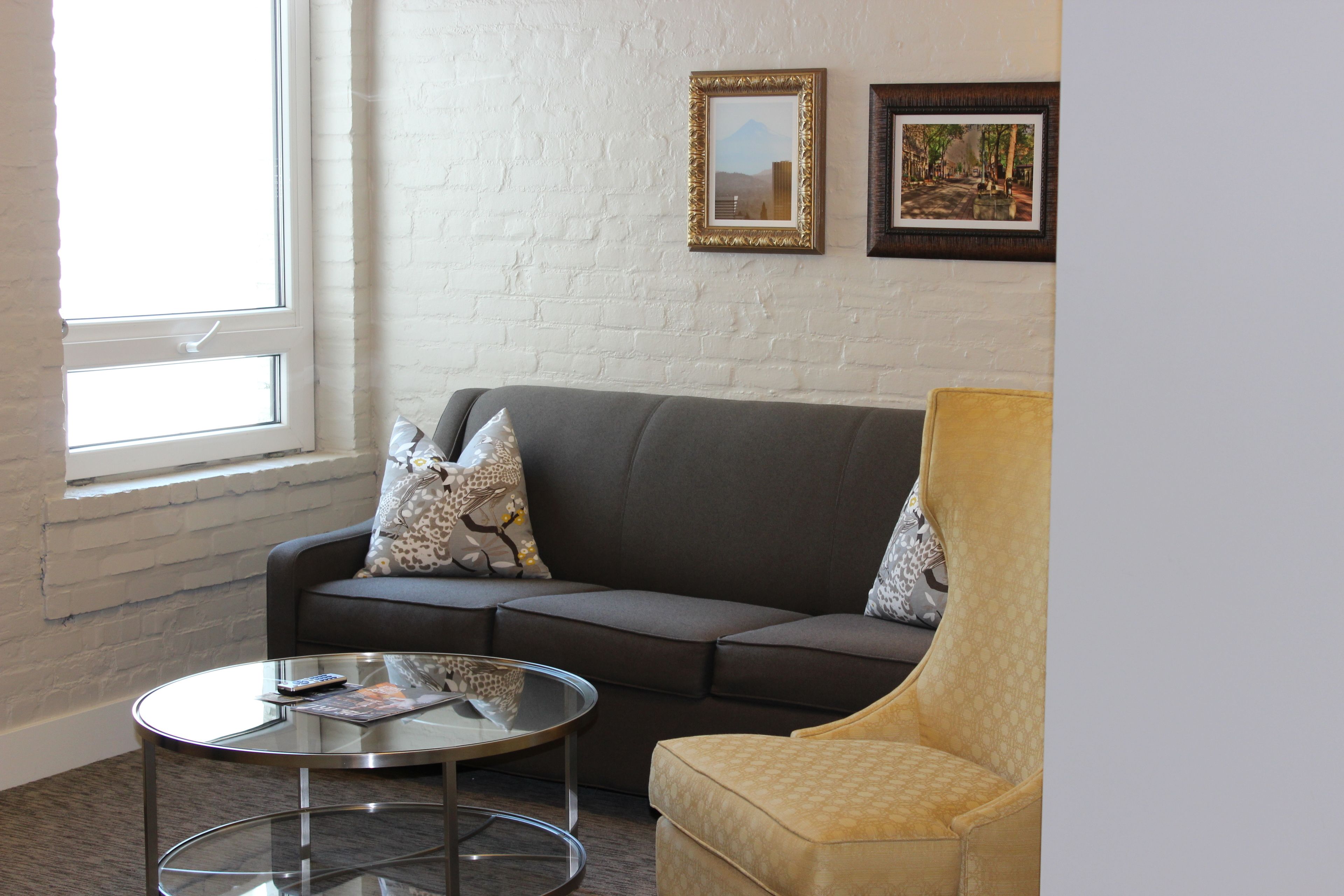 A living room with a grey sofa, a yellow armchair, a glass coffee table, and two framed pictures on the wall.