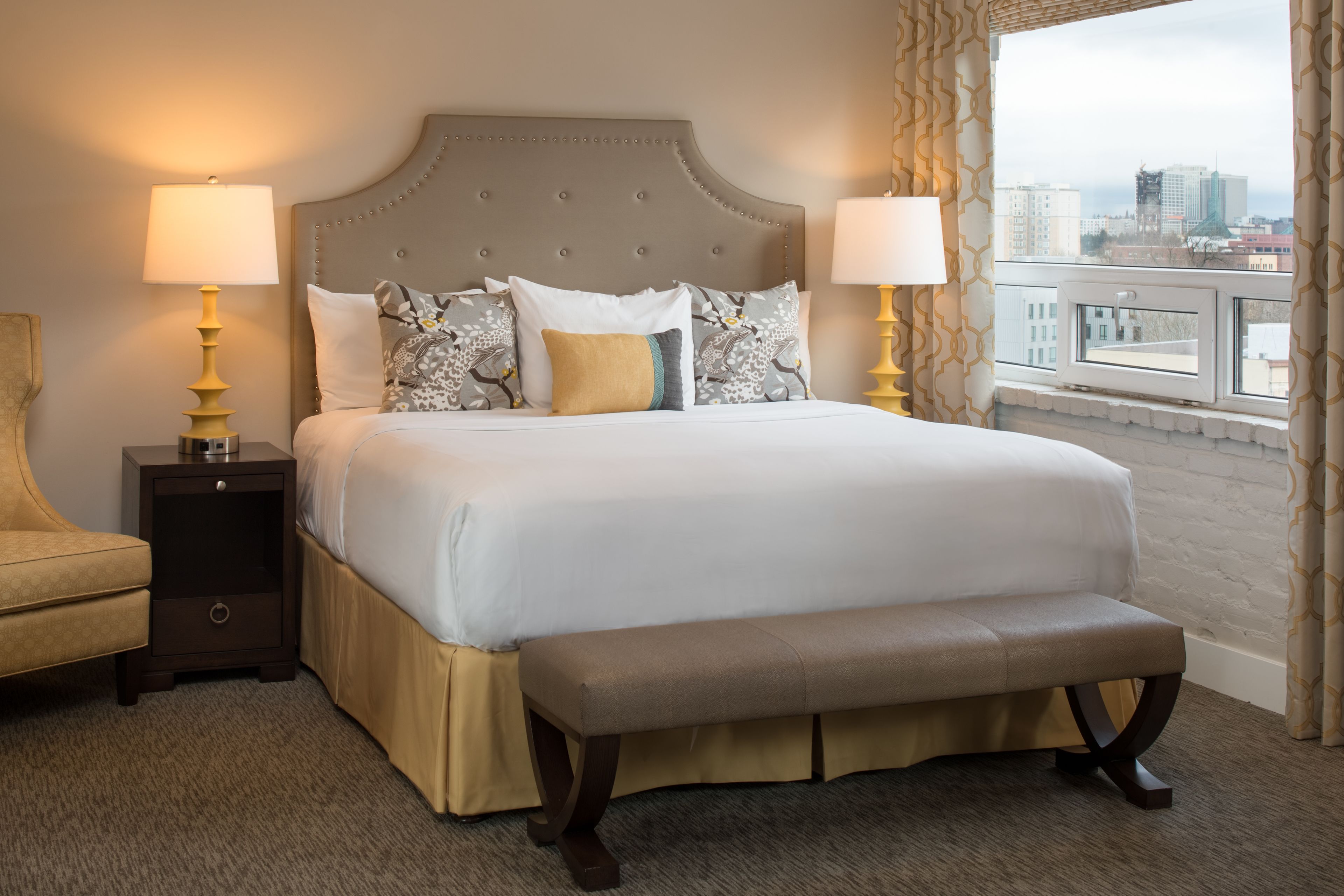 A hotel room with a large bed, a bench, two bedside tables with lamps, a chair, and a view of the city through a window.