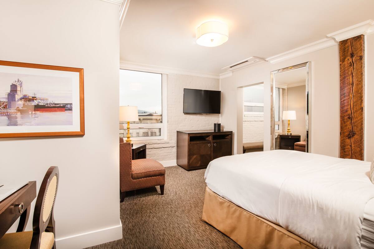 A hotel room with a bed, a desk with a laptop, a chair, a TV mounted on the wall, and a framed picture on the wall.