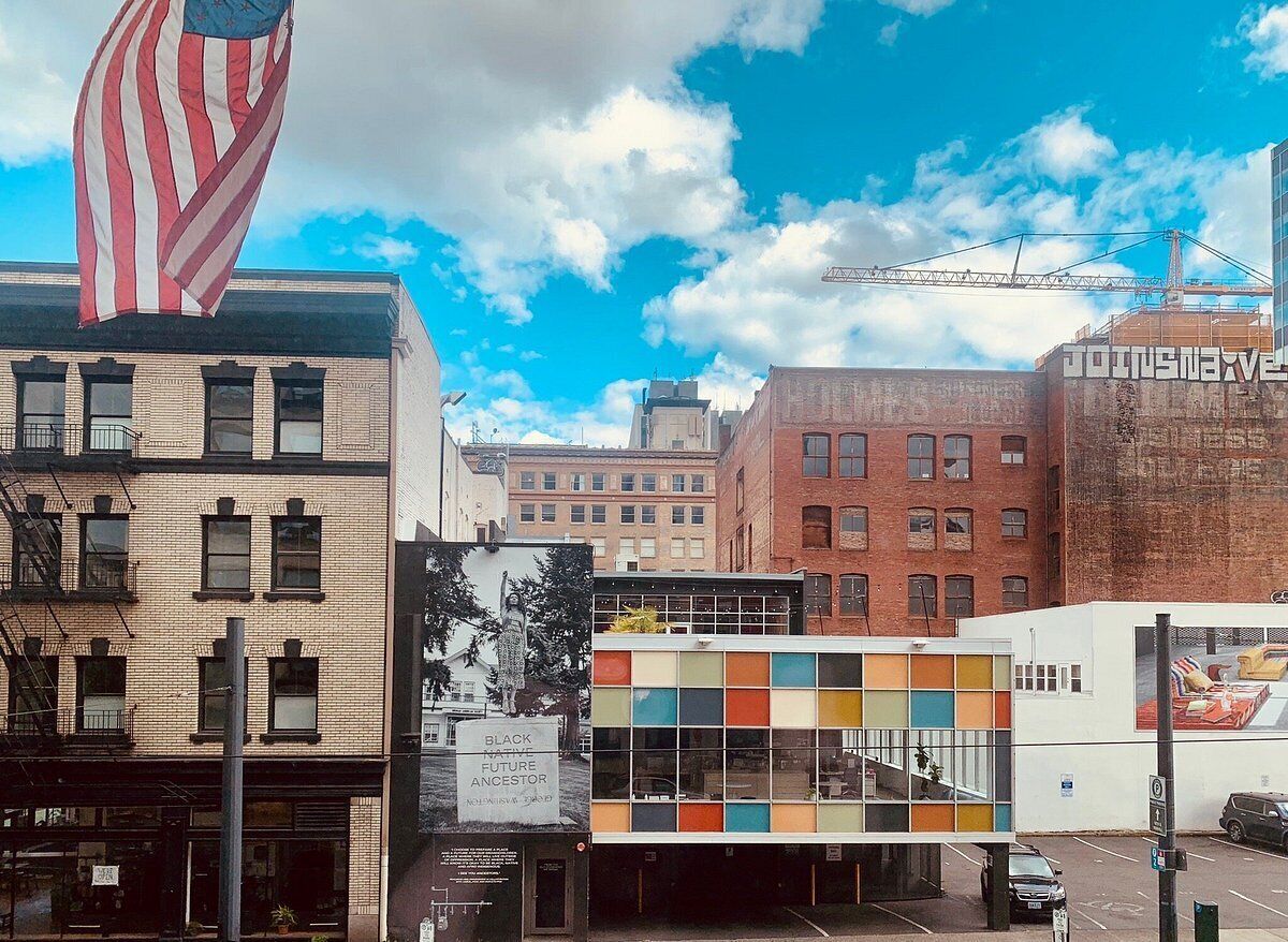 A multi-story building with a mix of brick and modern glass facades, a large American flag, and a crane in the background.
