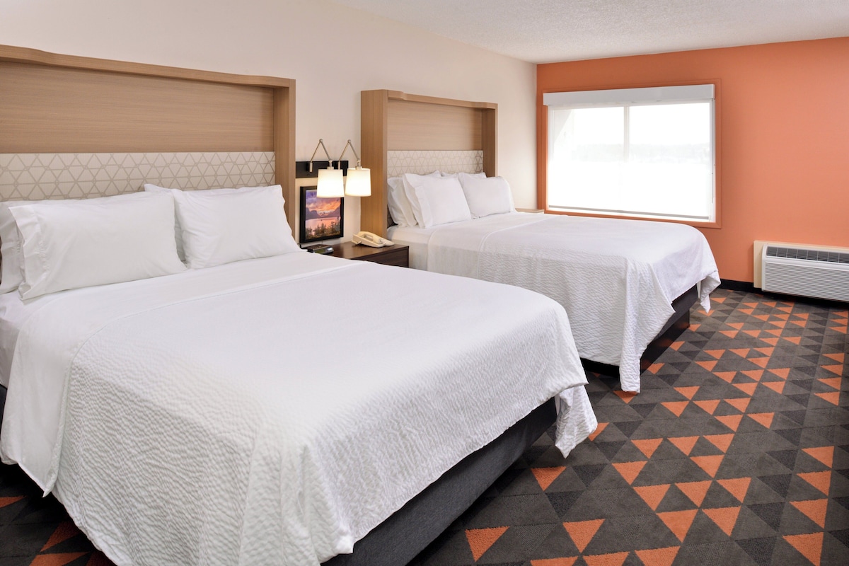 A hotel room with two beds, a nightstand with a lamp, a phone, and a window with blinds.
