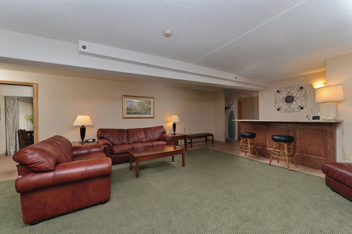 A living room with a brown leather sofa, a wooden coffee table, and two lamps. There is a painting on the wall and a bar area with stools.