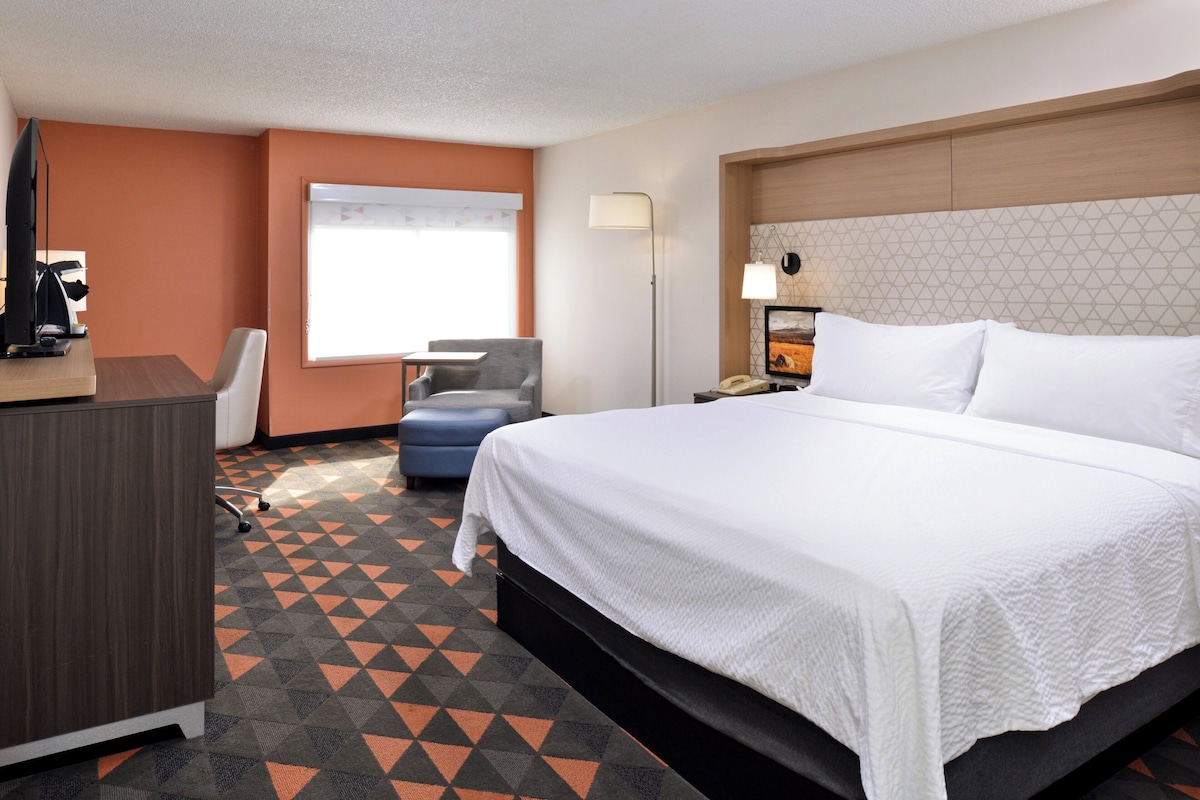 A hotel room with a large bed, a desk, a chair, a desk lamp, and a window with curtains.