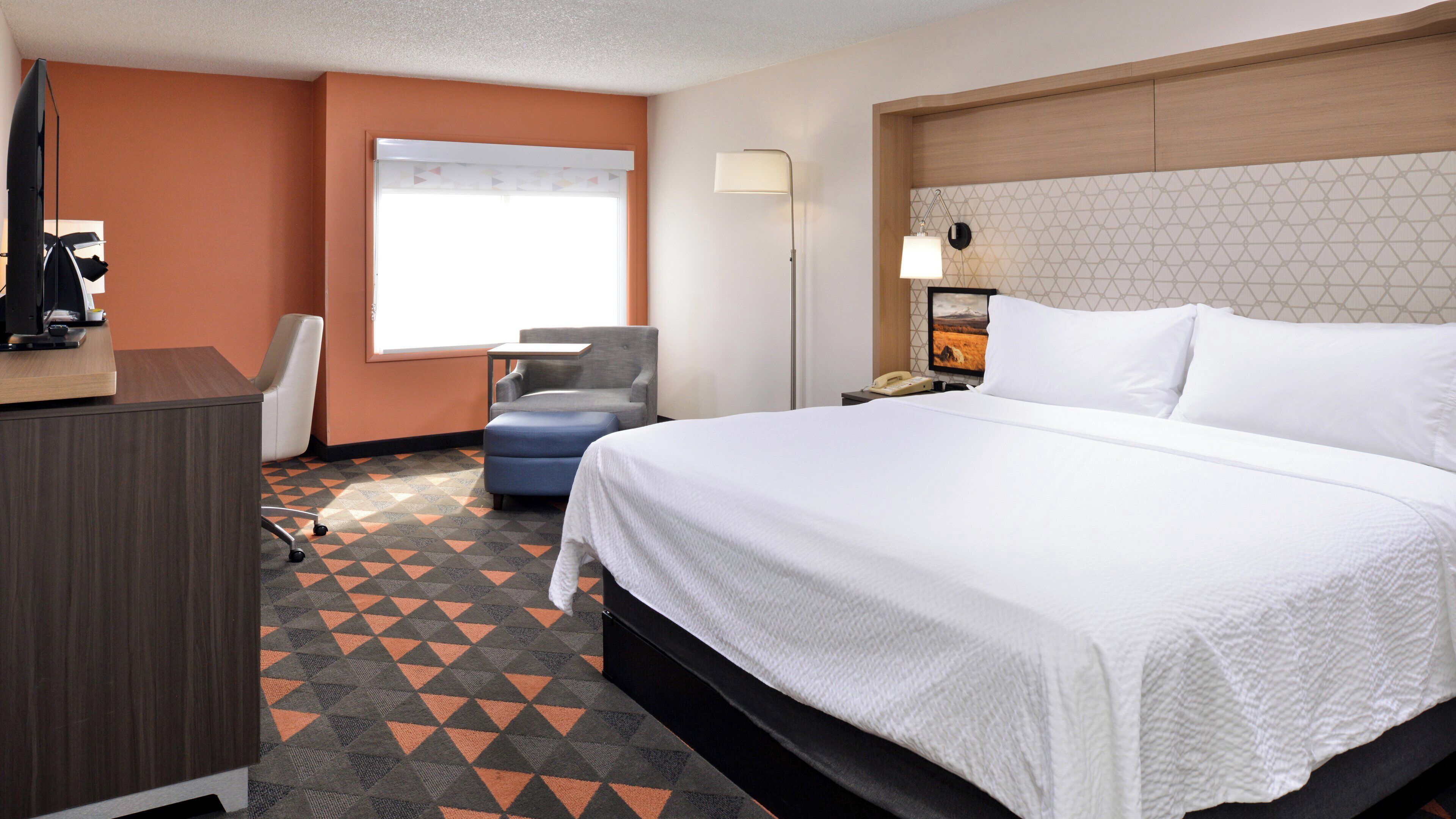 A hotel room with a bed, desk, chair, and a window with blinds.