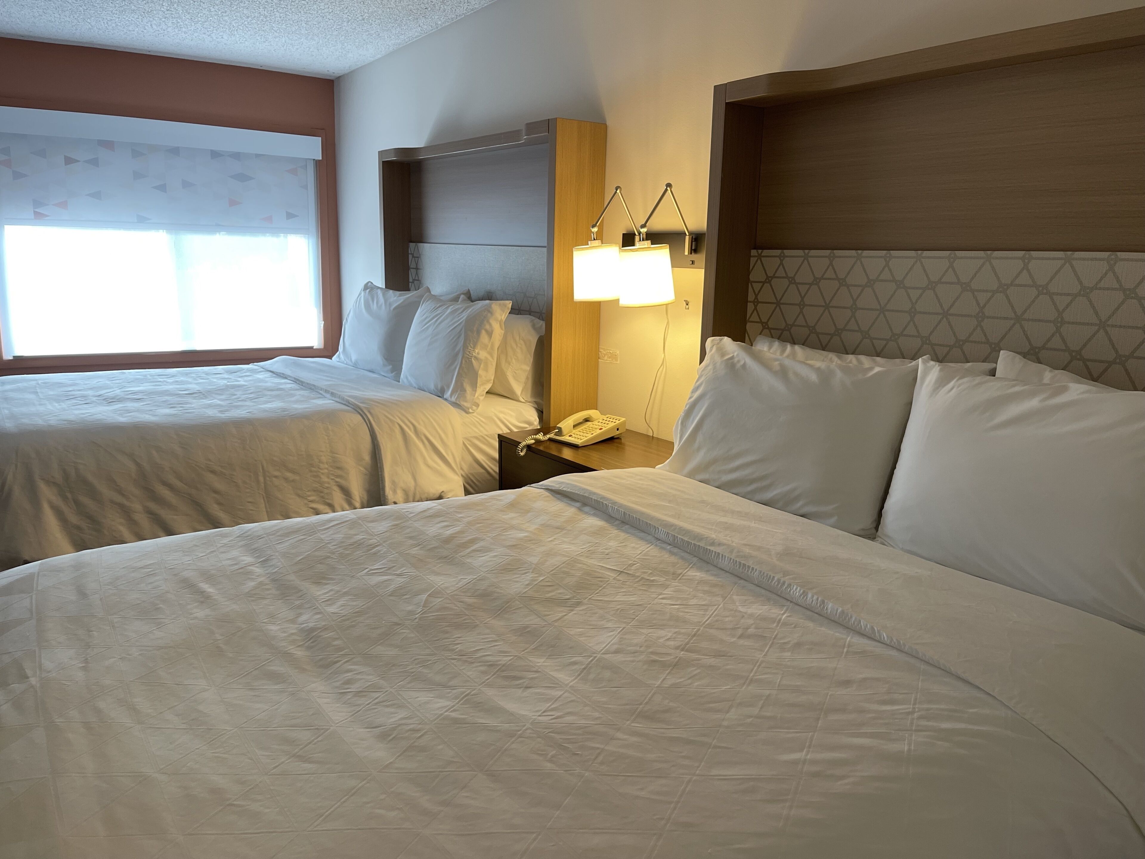 A hotel room with two beds, a nightstand with a phone, and a wall-mounted lamp.