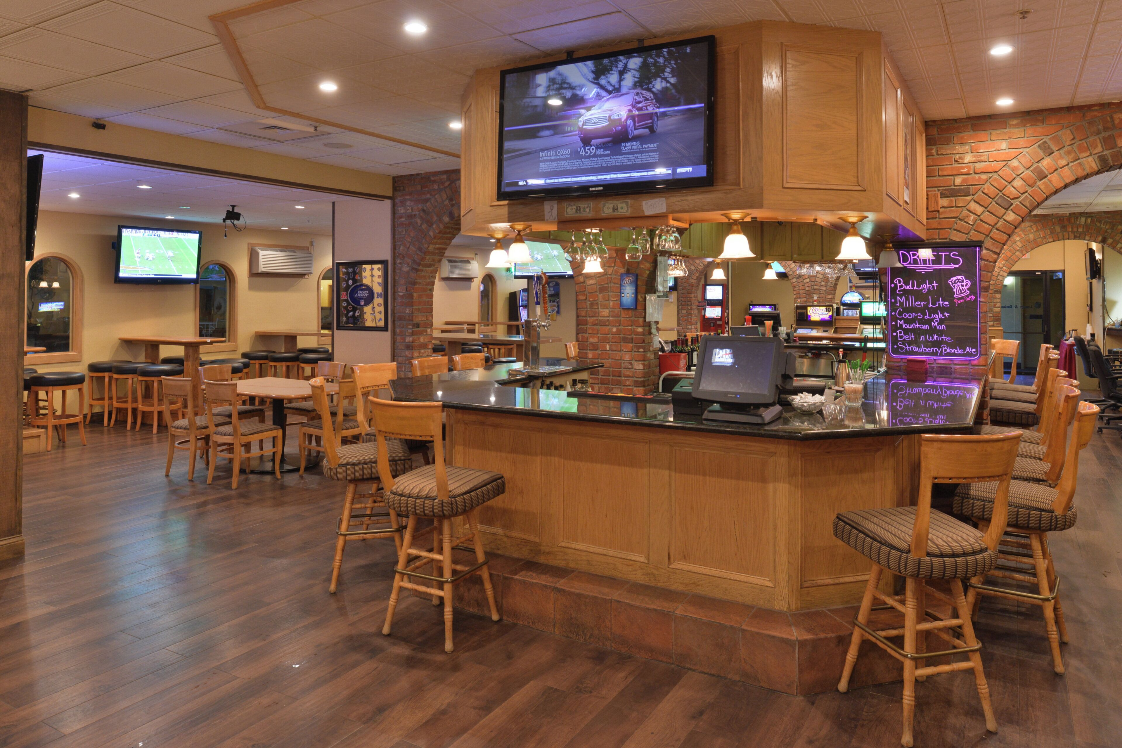 A bar area with wooden furniture, a television mounted on the wall, and a neon sign listing drink options.