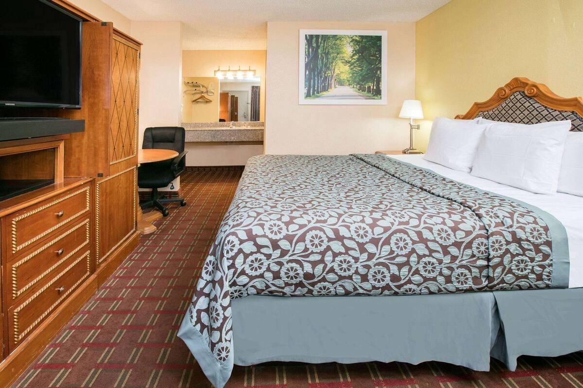 A hotel room with a bed, a TV, a desk, a chair, a mirror, and a painting on the wall.