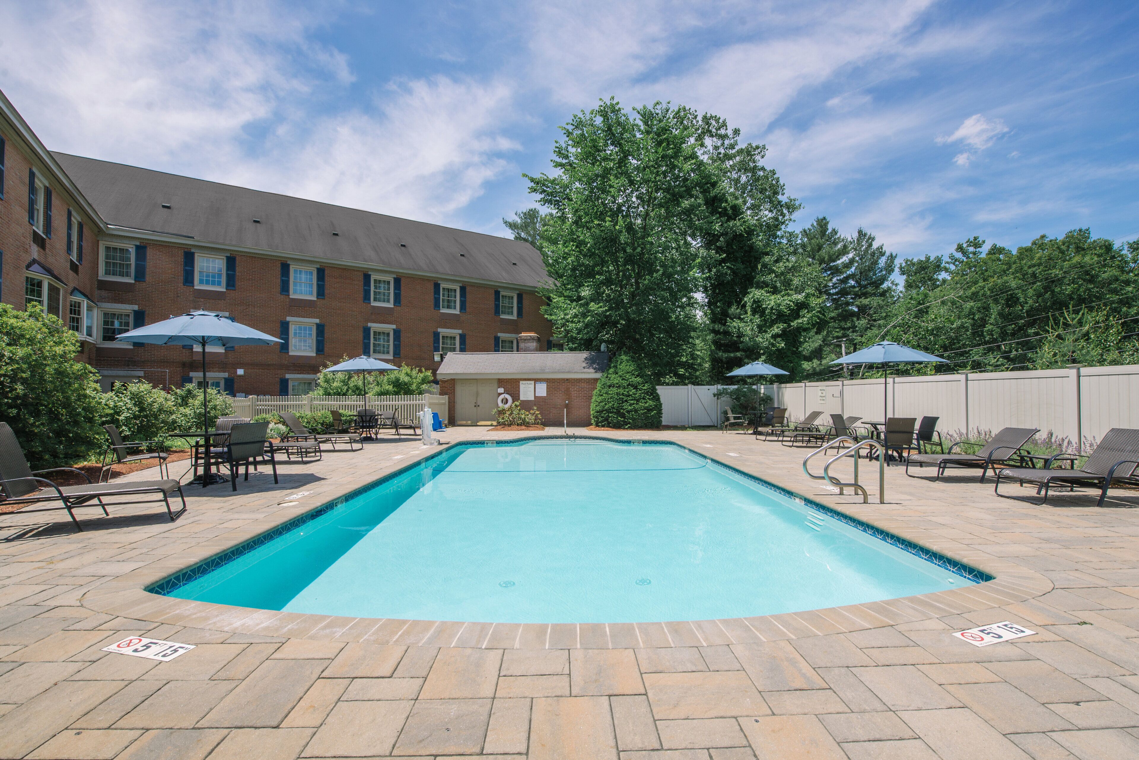 A rectangular swimming pool with a tiled edge, surrounded by lounge chairs and umbrellas, with a brick building in the background.