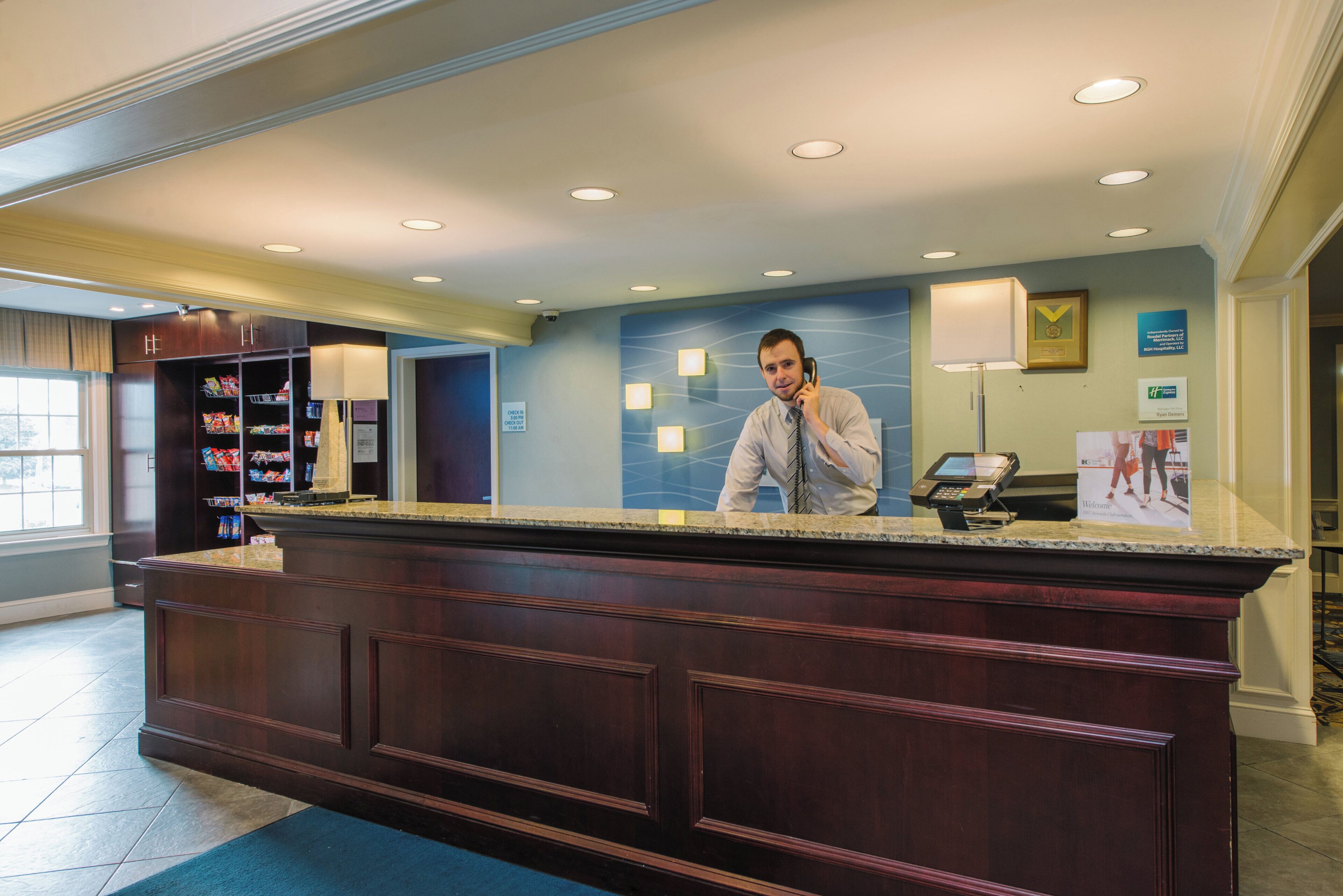 Een receptiebalie in een hotel met een incheckbalie, een beveiligingscamera, een telefoon bevestigd aan de muur en een ingelijste onderscheiding aan de wand.