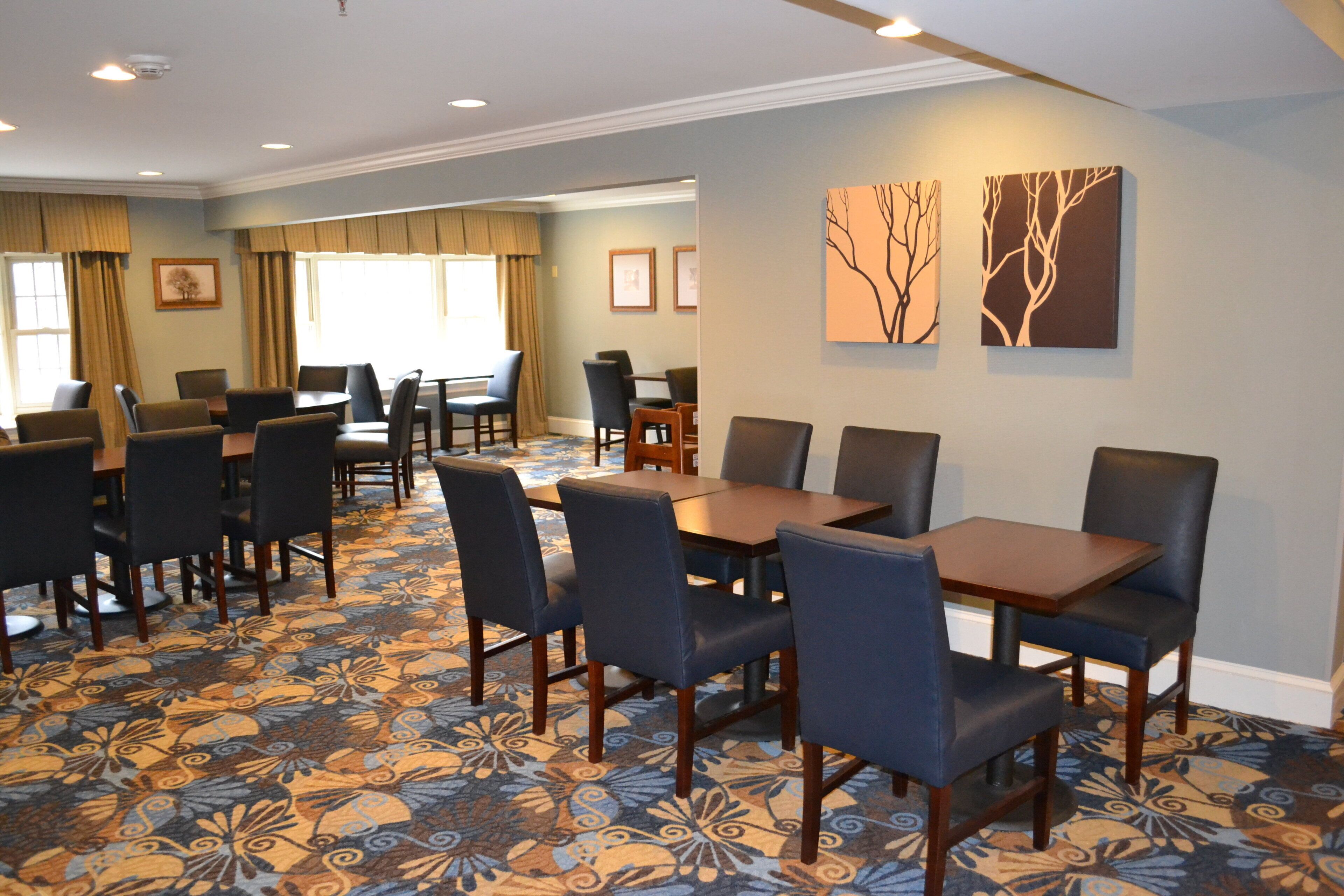 A hotel dining area with a patterned carpet, multiple tables and chairs, and artwork on the walls.