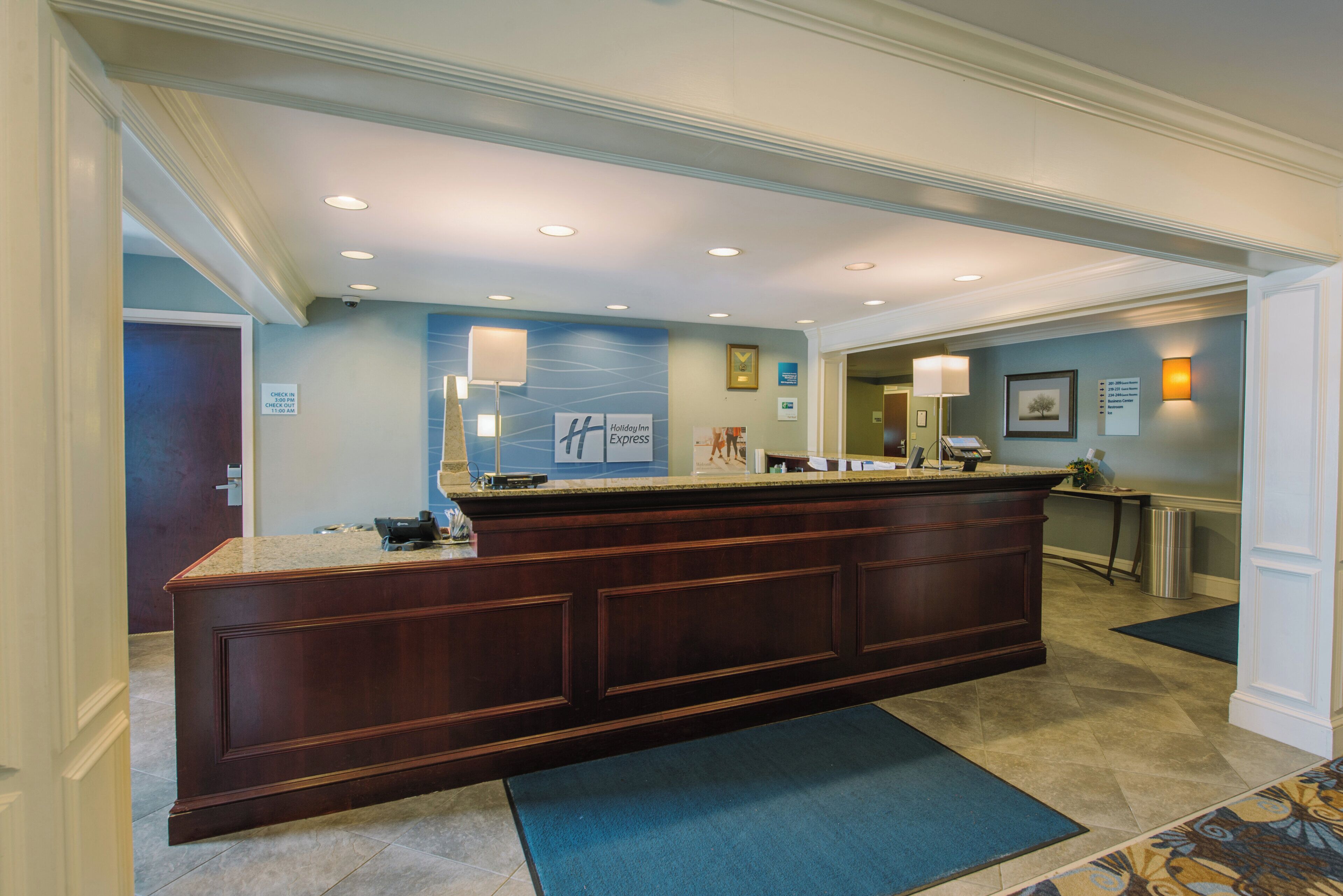 A hotel lobby with a reception desk, a blue carpet, and a wall with various signs and a framed picture.