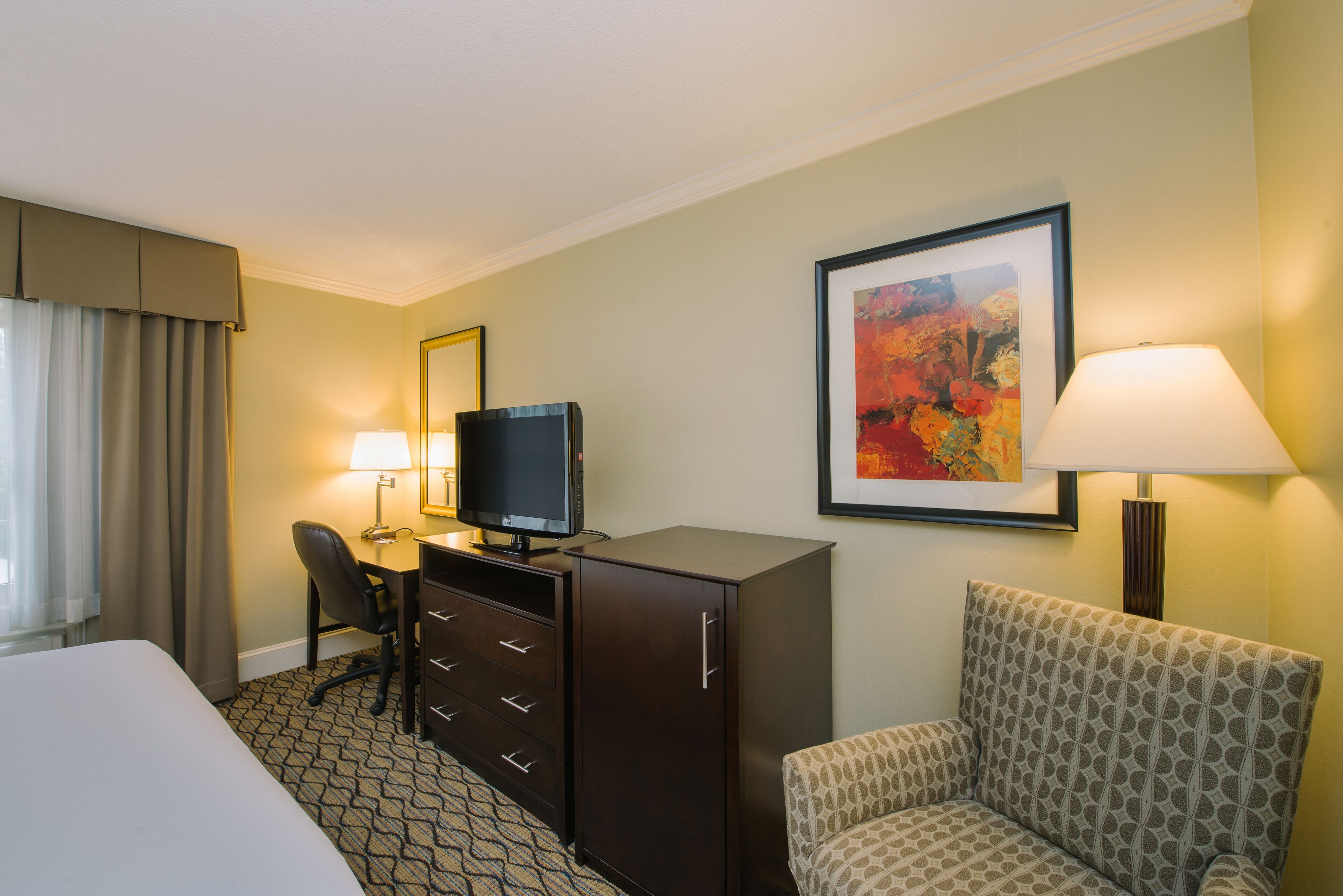 Una habitación de hotel con una cama, un escritorio con televisor, una silla, un aparador, una lámpara y un cuadro en la pared.