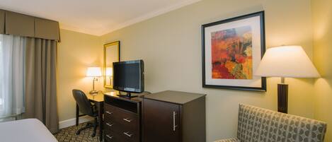 A hotel room with a bed, a desk with a television, a chair, a dresser, a lamp, and a framed picture on the wall.