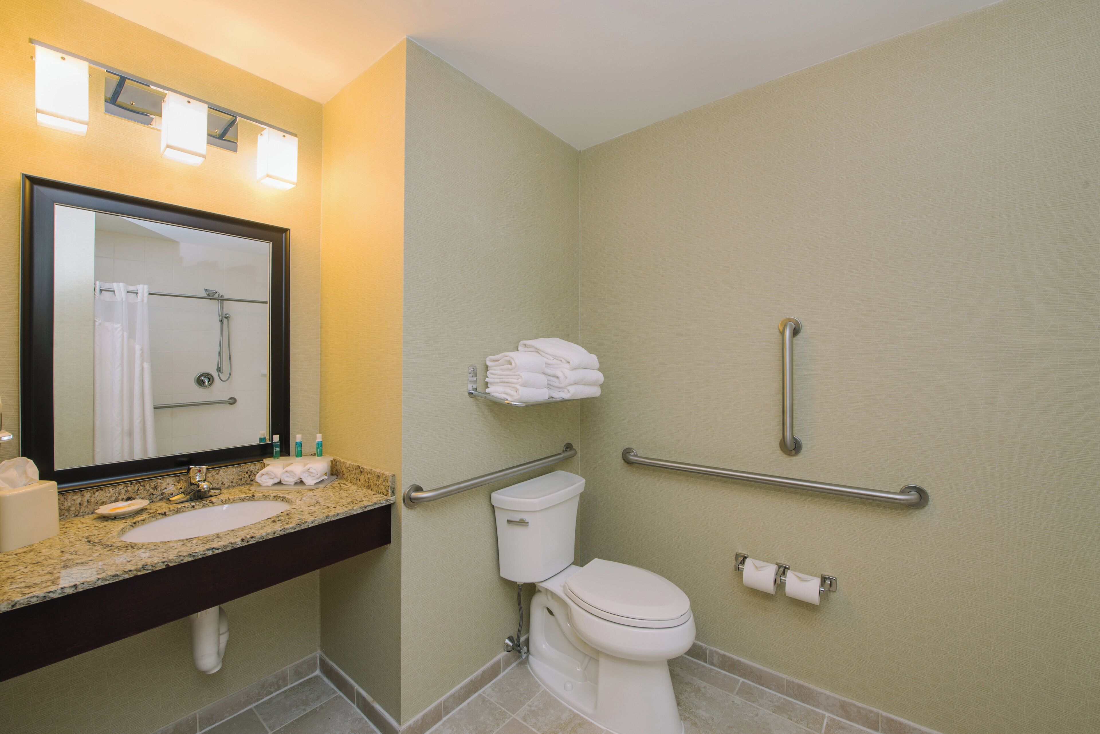 A bathroom with a sink, mirror, toilet, grab bars, and a towel rack.