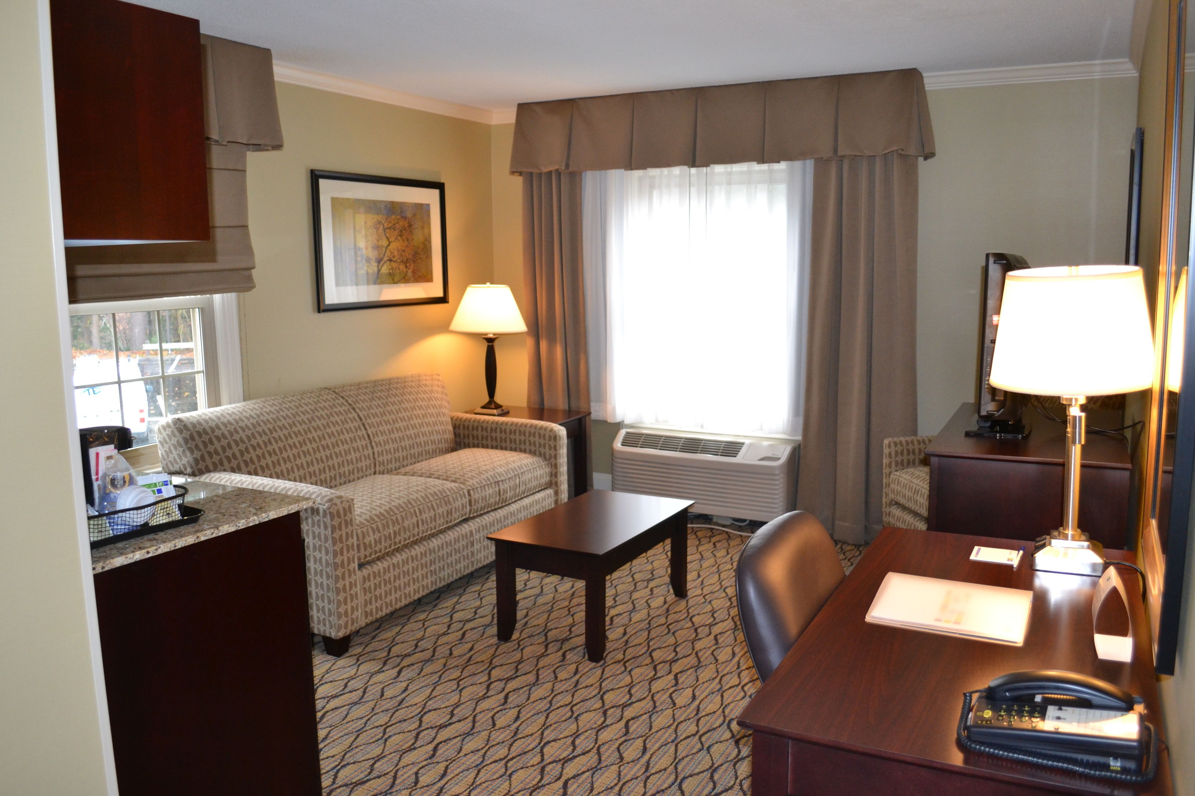 A hotel room with a patterned sofa, a small table, a desk with a lamp, a telephone, and a window with curtains.