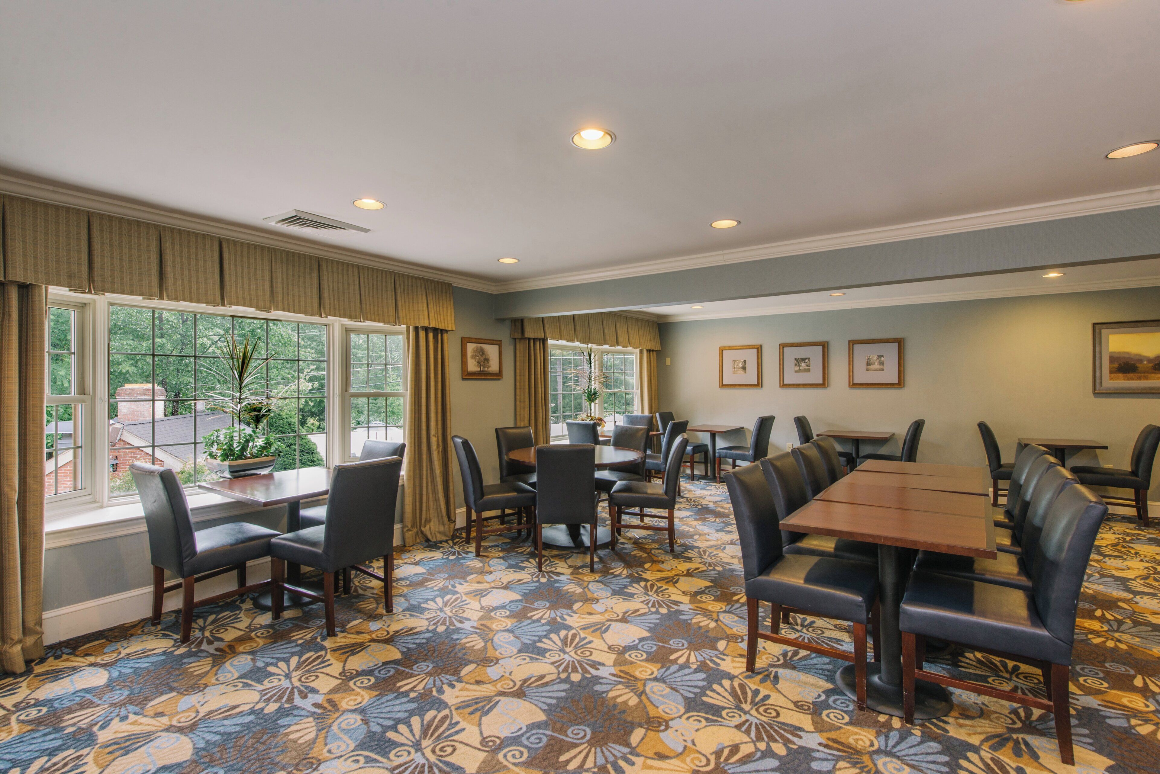 A hotel room with a large dining table, multiple chairs, and a patterned carpet.