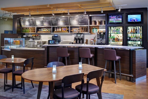 A bar area with wooden tables and chairs, a counter with a menu board, and a refrigerator stocked with bottles.