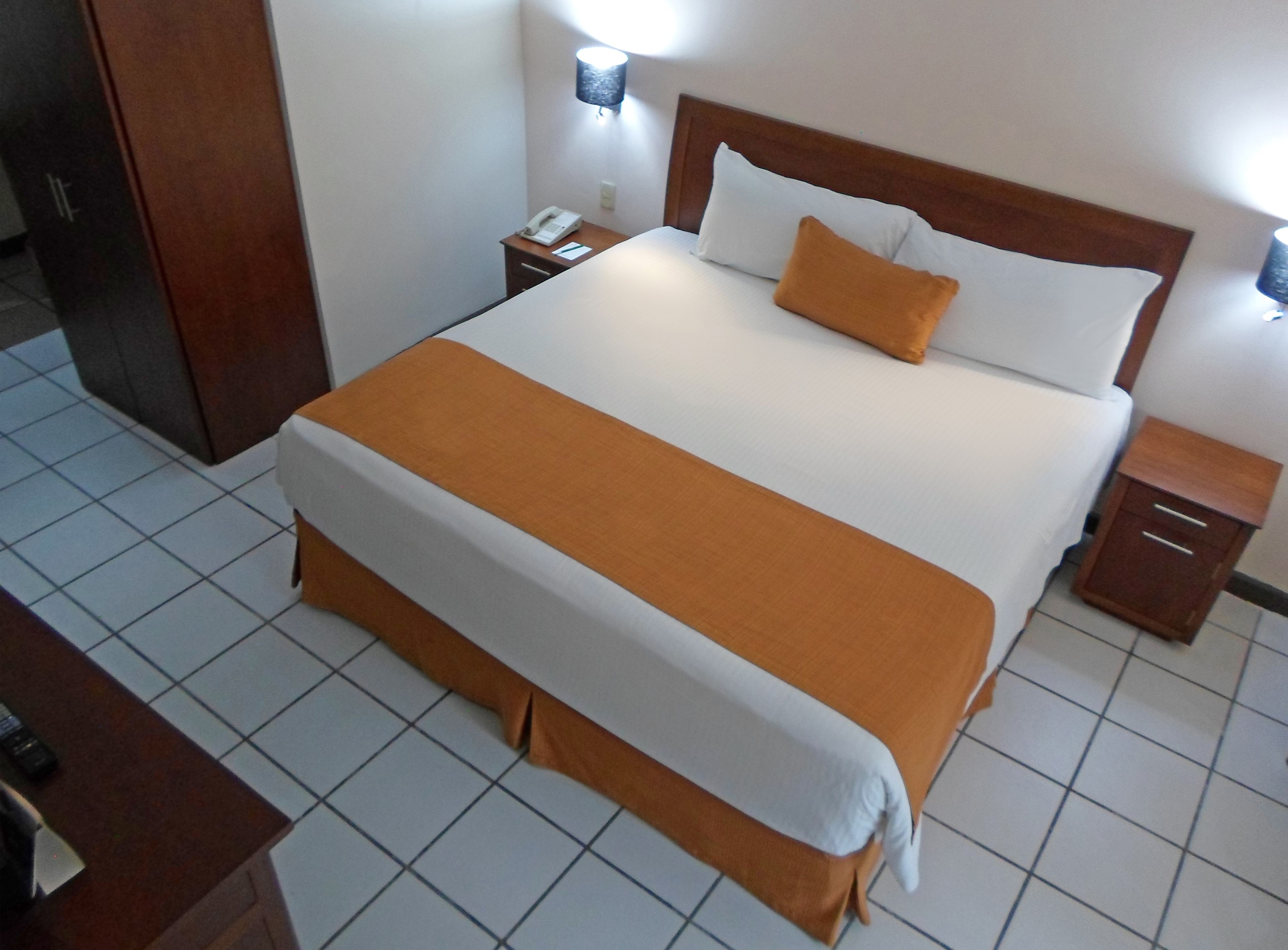 A hotel room with a large bed, a bedside table, a nightstand, and a wall-mounted lamp.