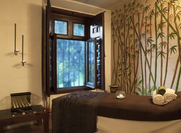 Quarto com cama, janela com vista para árvores, parede com padrão de bambu e uma pequena mesa com velas.