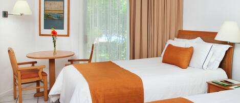 A hotel room with two beds, a small table with a chair, a lamp, and a painting on the wall.