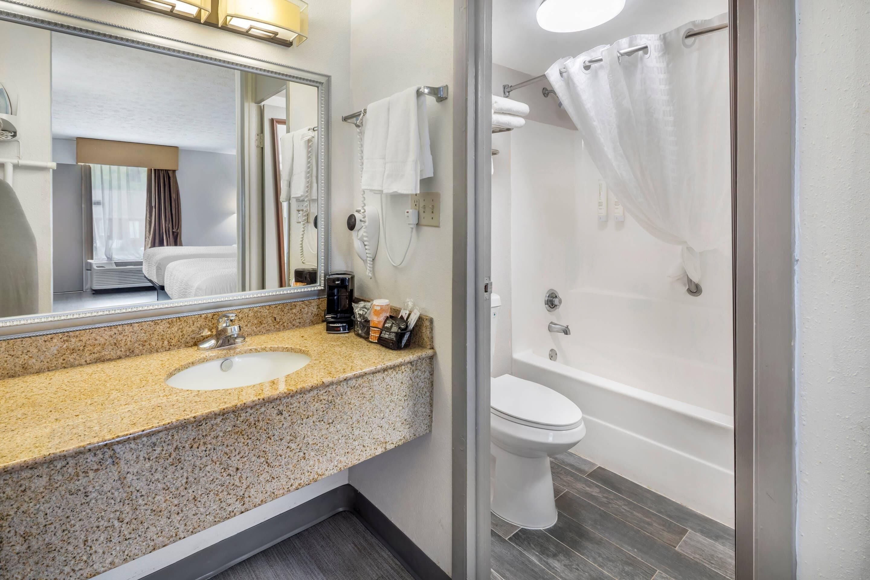 Une salle de bain d’hôtel avec un lavabo au comptoir en granit, un miroir, des toilettes blanches et une baignoire avec un rideau de douche.