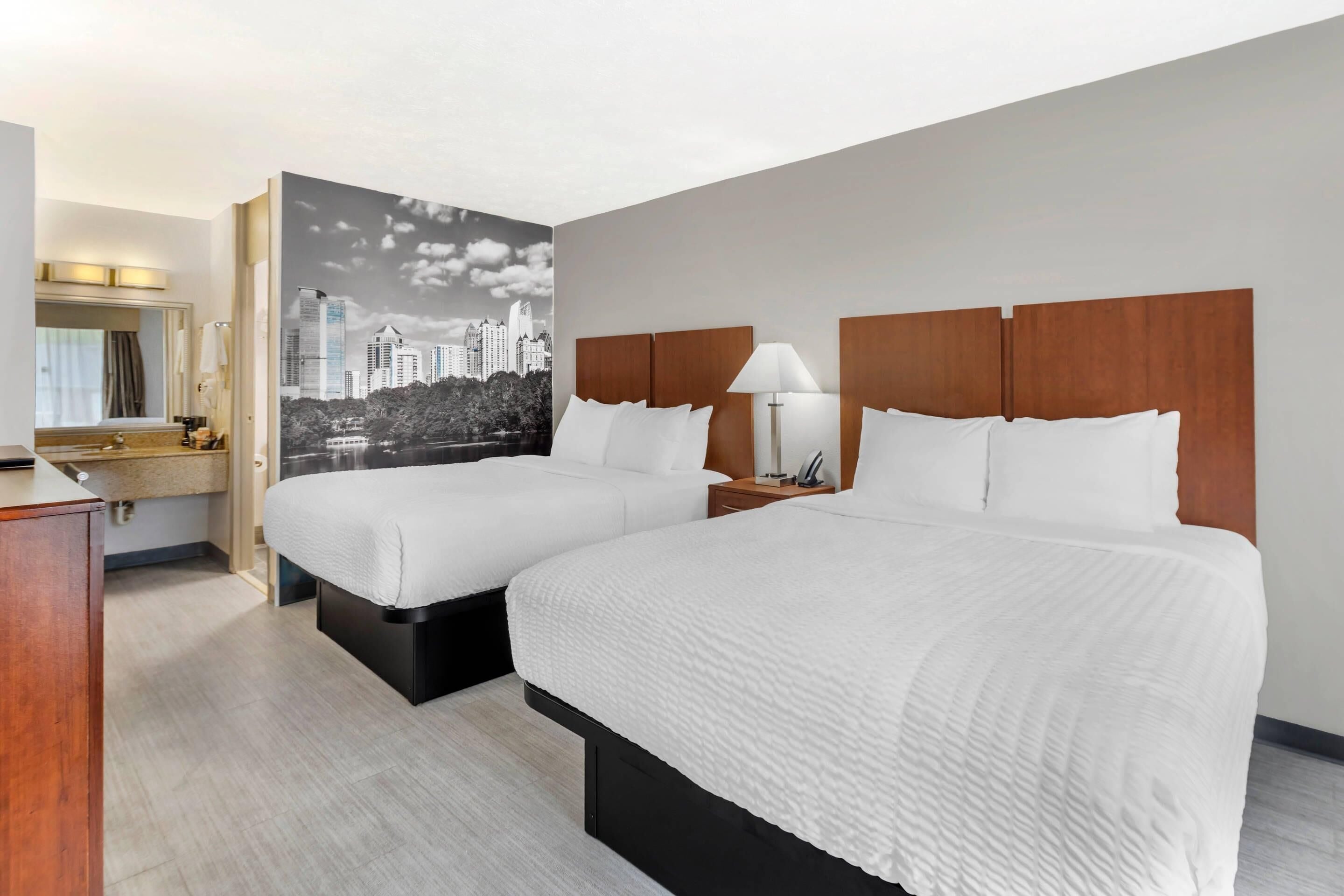 Une chambre d’hôtel avec deux lits, un grand miroir, une décoration murale représentant un paysage urbain et une table de chevet en bois avec une lampe.