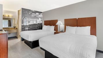 Une chambre d’hôtel avec deux lits, un grand miroir, une décoration murale représentant un paysage urbain et une table de chevet en bois avec une lampe.