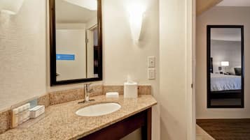 Casa de banho de hotel com lavatório de bancada em granito, espelho, dispensador de toalhas e porta visível que dá para um quarto com cama e mesa de cabeceira.