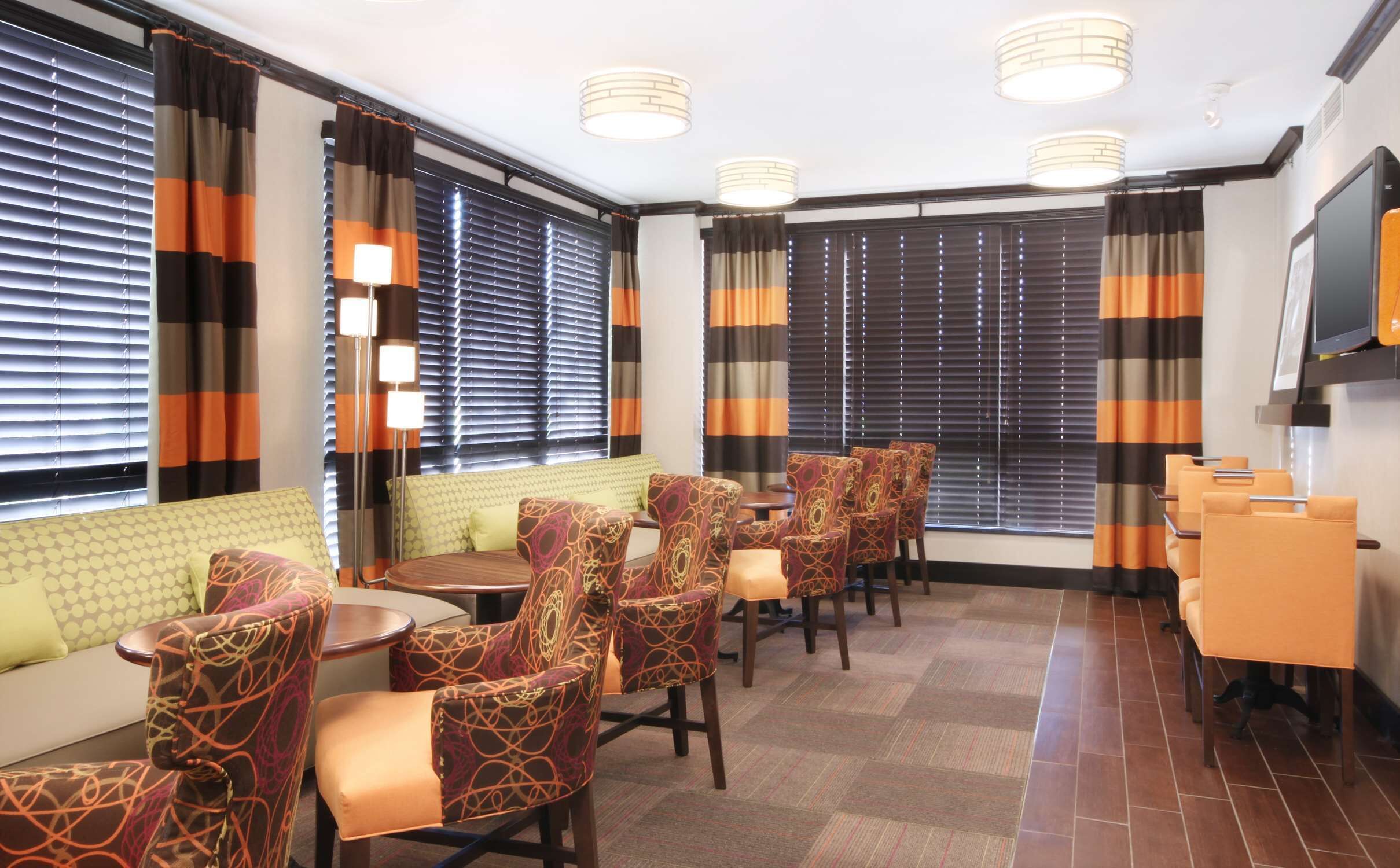 Habitación de hotel con zona de estar que incluye sillas estampadas y un sofá, ventanas grandes con persianas y un televisor de pantalla plana montado en la pared.