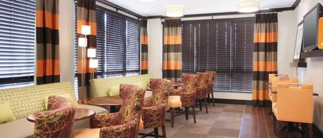 Habitación de hotel con zona de estar que incluye sillas estampadas y un sofá, ventanas grandes con persianas y un televisor de pantalla plana montado en la pared.